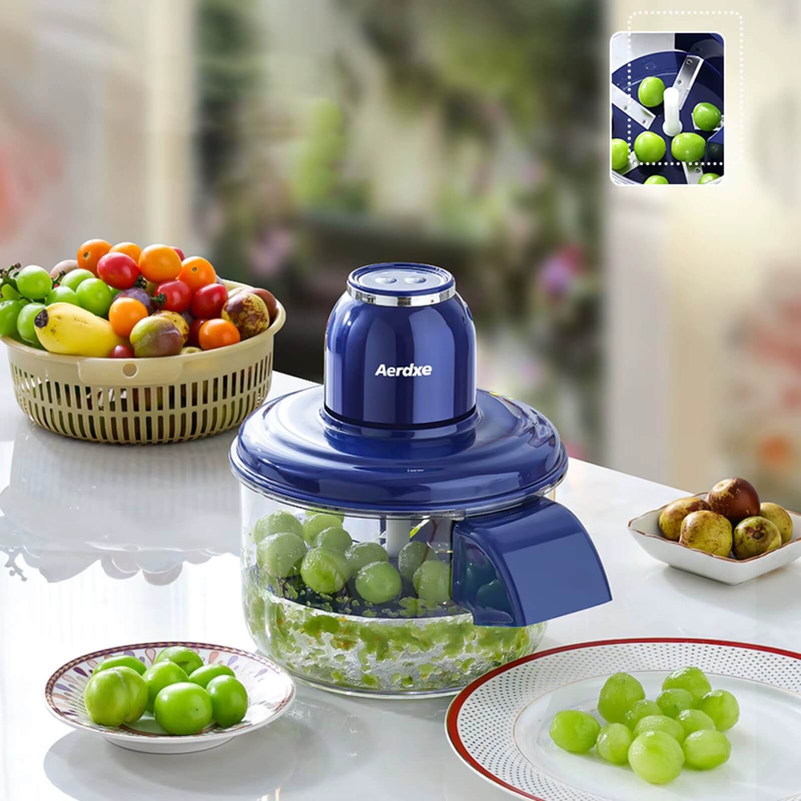Blue food chopper with grapes on a table, surrounded by fruits and vegetables.